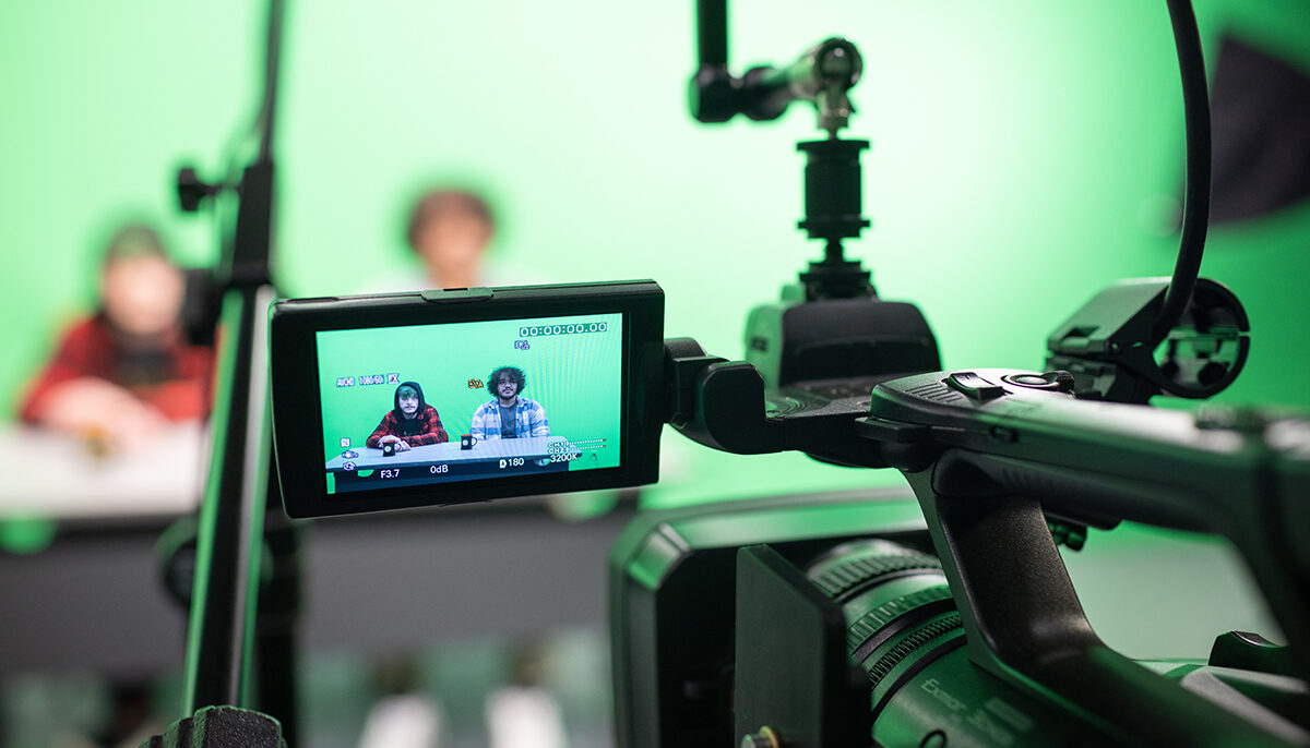 An LCD screen of a studio video camera is seen in focus. Two people can be seen sitting at a table through the screen. The rest of the image is blurred with a green tint.