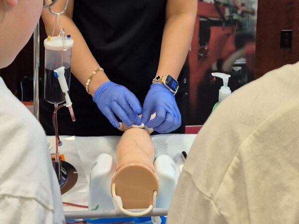 Two students in white t-shirts observe a medical simulation demonstration. An instructor wearing blue nitrile gloves and a smartwatch works on a realistic training model of a human forearm, applying gauze or a bandage. Beside the arm model is an IV bag filled with liquid hanging on a stand, connected to medical tubing.