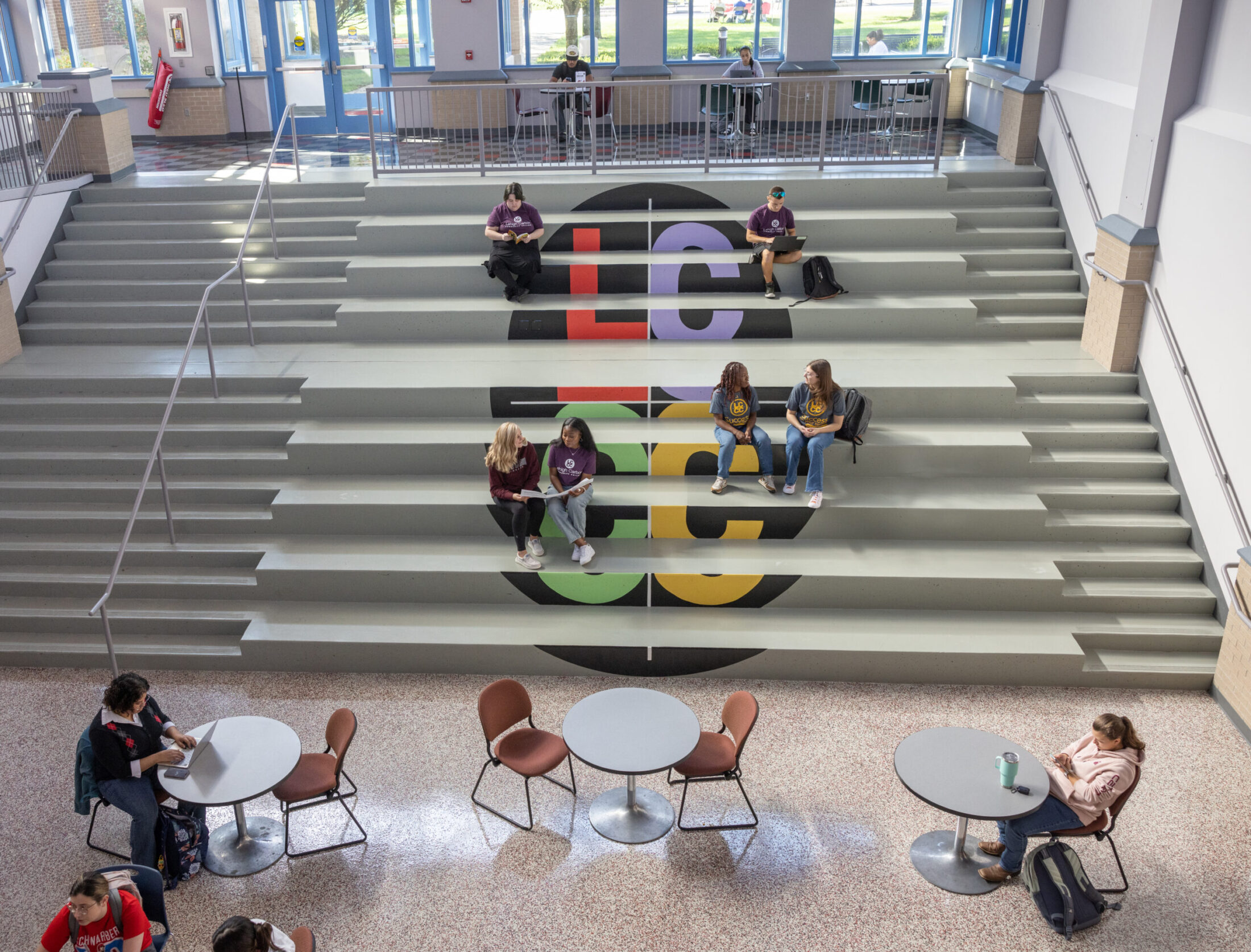 An overhead view of students sitting and studying in a bright, modern common area at Lehigh Carbon Community College, featuring a large set of tiered steps painted with the LCCC logo.