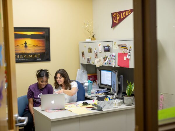 An office setting at Lehigh Carbon Community College (LCCC) where a staff member or advisor is sitting at a desk and showing information on a laptop screen to a student. The room features a desk with a computer monitor, a poster on the wall, and a pennant. The photo is framed as if looking through an open doorway.