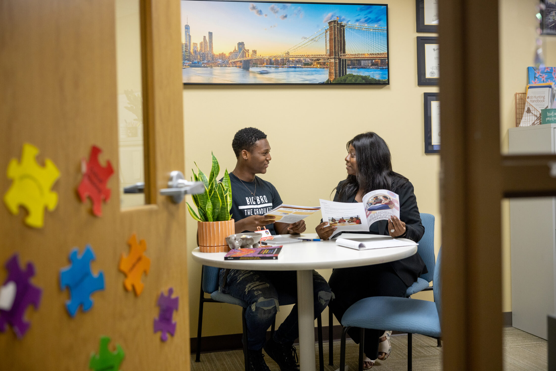 An office setting at Lehigh Carbon Community College (LCCC) where two individuals are seated at a round table, engaged in a conversation or advising session. The wall to the left features colorful puzzle piece decorations, and a large photo in the background shows a city skyline.