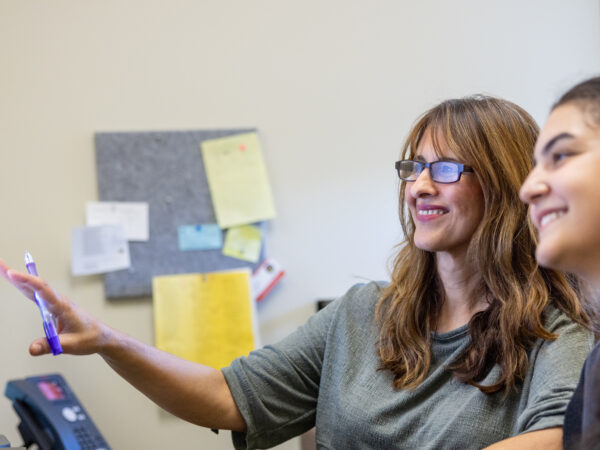 An older female advisor with glasses pointing a pen toward a computer monitor while sitting next to a young female student looking at the screen in an office.