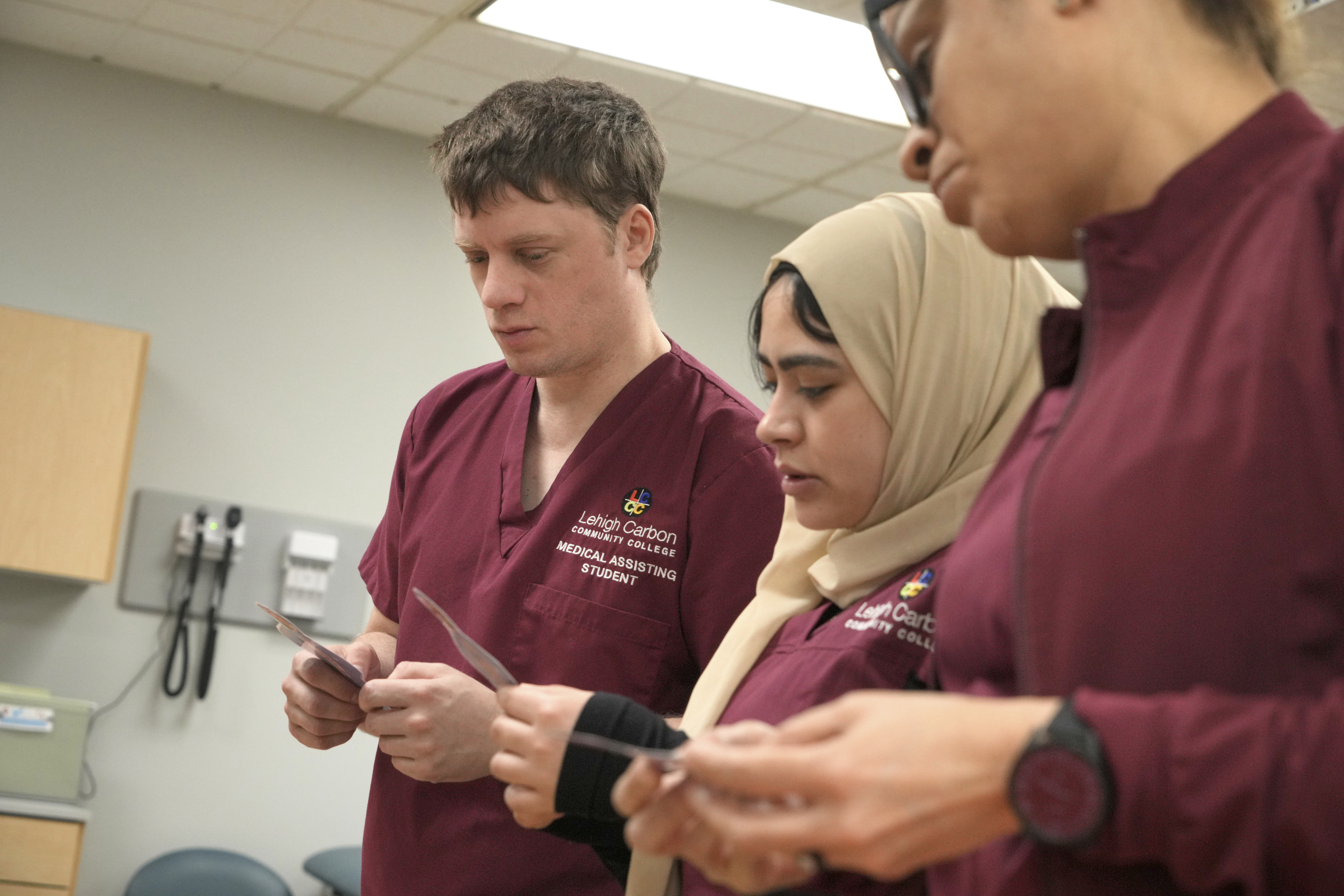 **Alt Text:** Three Lehigh Carbon Community College medical assisting students in maroon scrubs reviewing patient cards or charts together in a clinical setting.