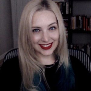 A blonde woman wearing red lipstick and a black shirt smiles at the camera.