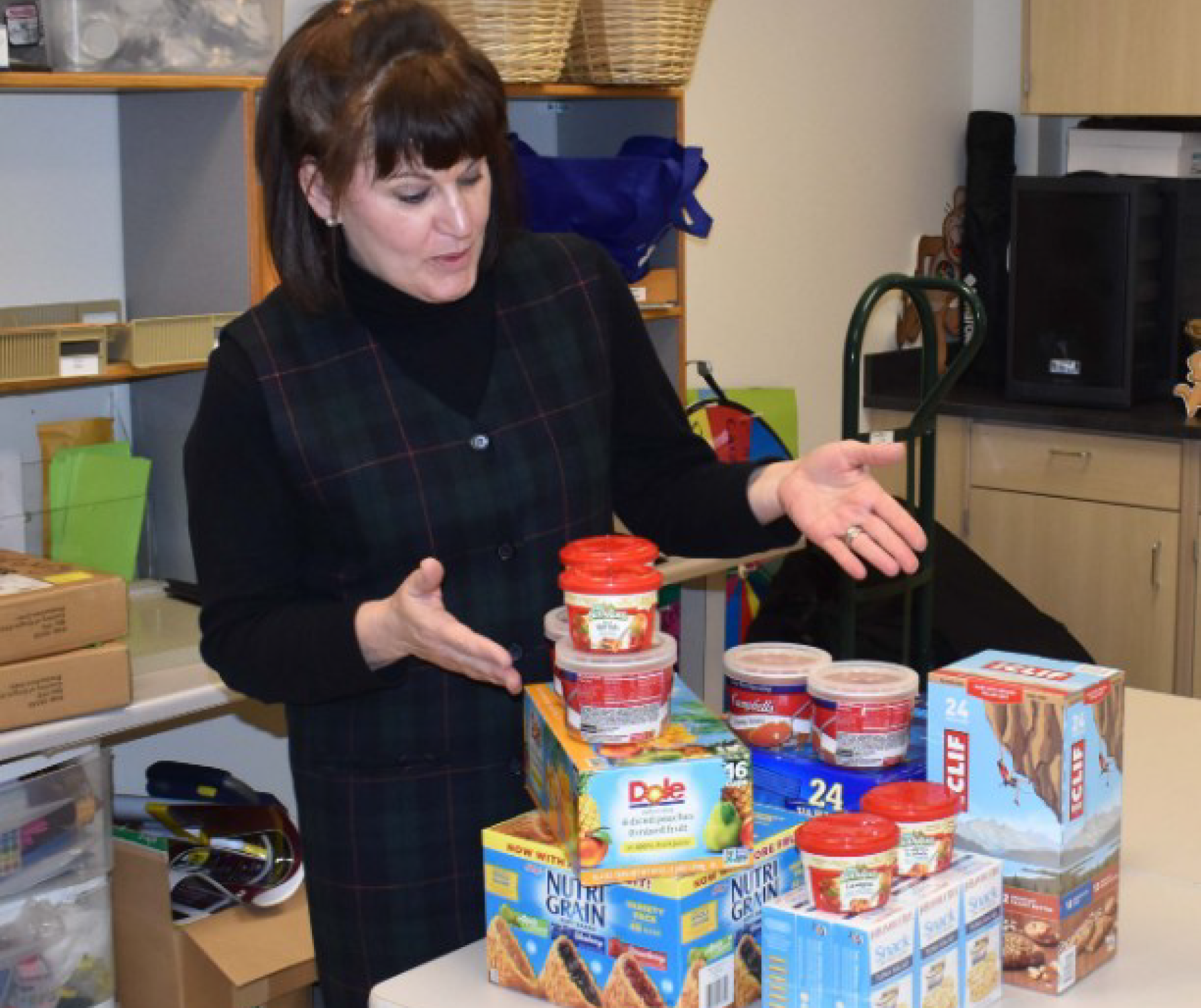 A woman with short dark brown hair gestures to a pile of pantry goods on a table.