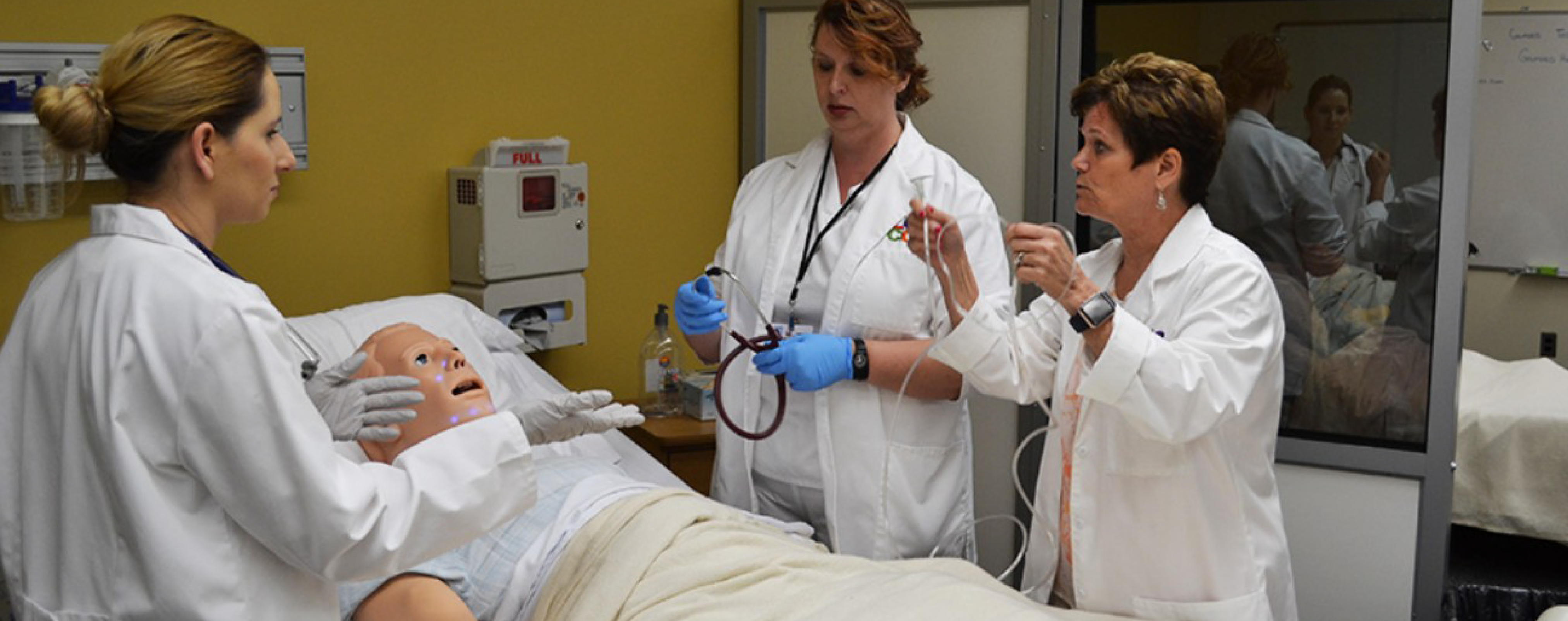 Three nurses in white lab coats stand around a nursing manikin while one of the nurses provides instruction.