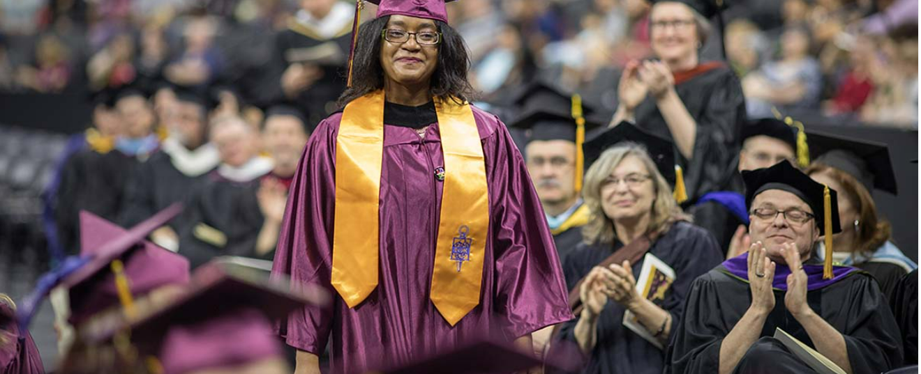 An African American woman wearing a maroon graduation cap and gown walks down a center aisle at graduation while faculty clap.