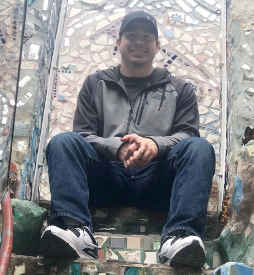 A man in a sweatshirt, jeans and ballcap, sits on a large concrete mosaic chair sculpture.