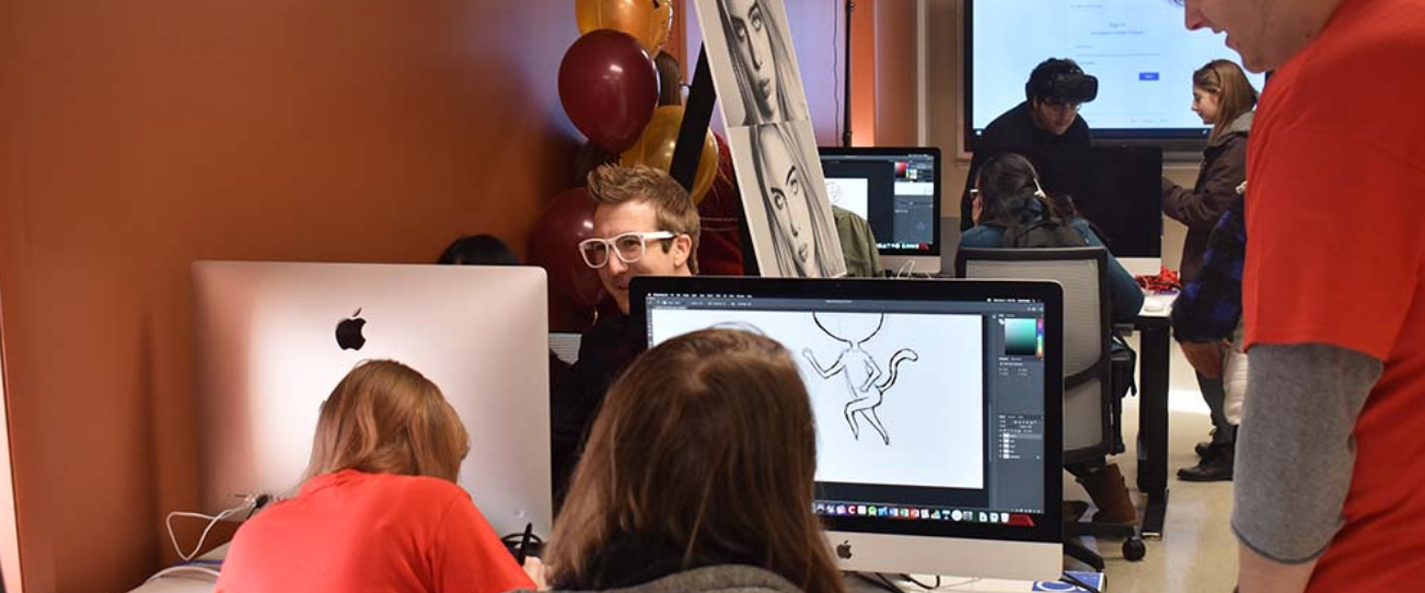 The backs of two students can be seen working on large mac screens. A man with white glasses is smiling on the other side of the computers.