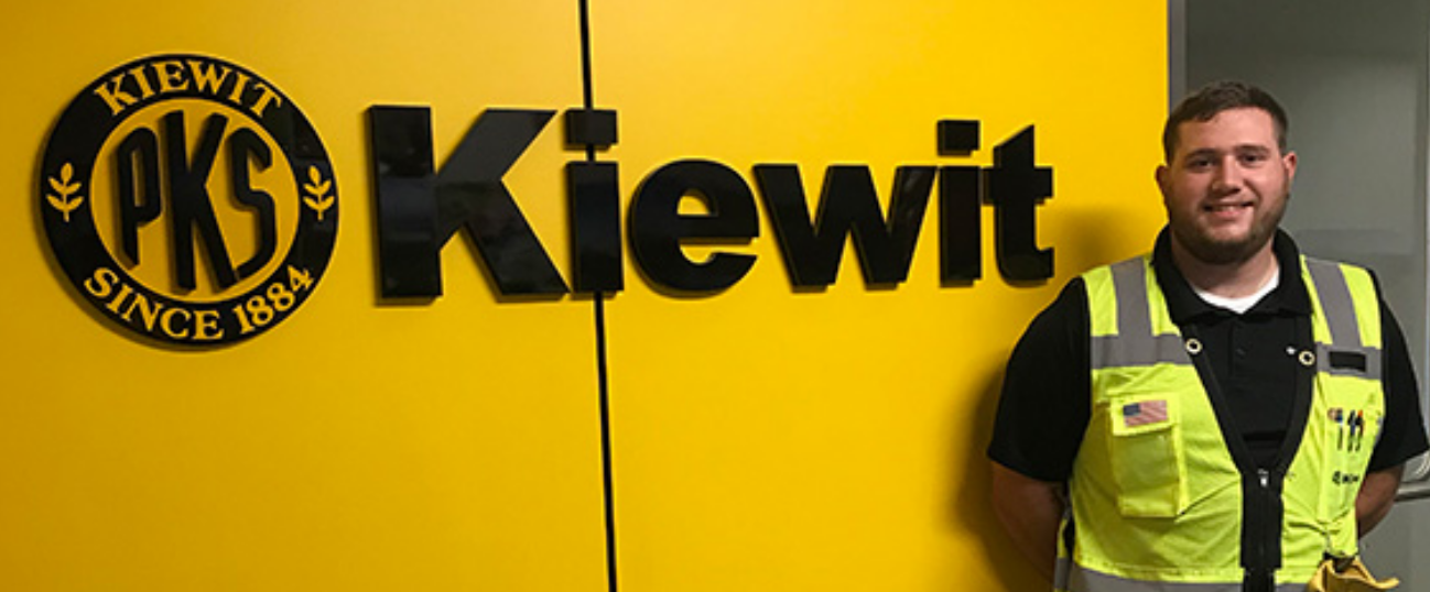 A man wearing a neon yellow high visibility vest stands against a yellow wall with black lettering that reads "Kiewit".