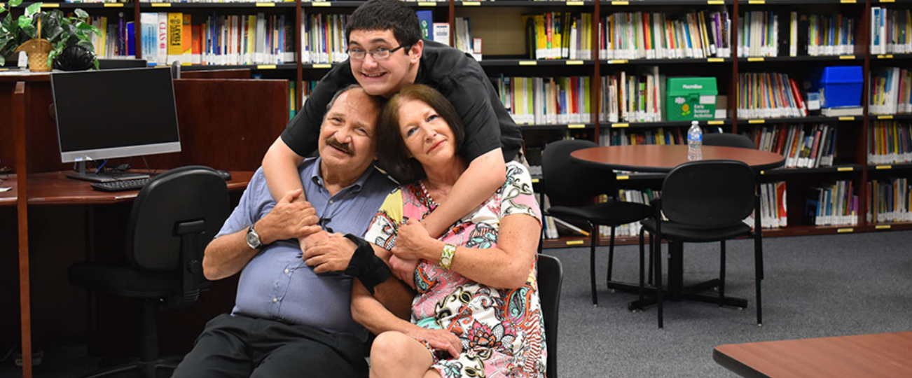 A student stands behind sitting parents and puts his arms around them in a room with a bookshelf in the background.