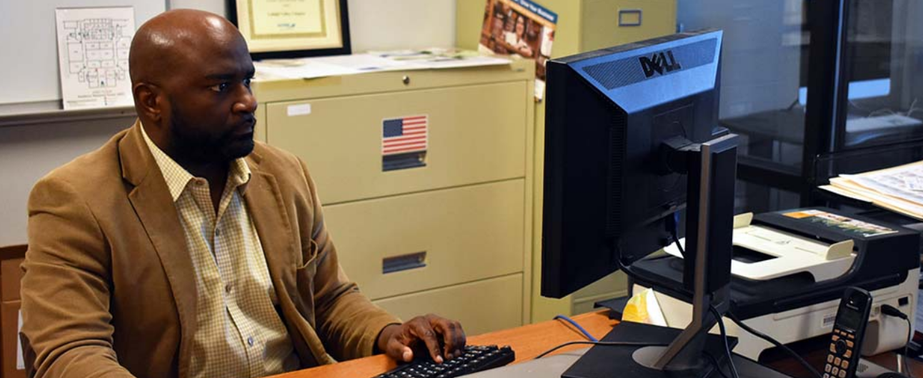 An African American man in a dress shirt and blazer sits in an office and works at a computer.