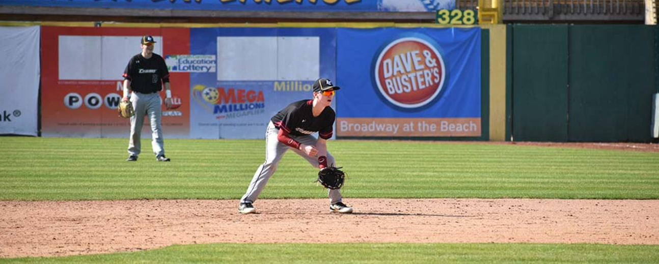A baseball player crouches on the field to catch a ball while another player stands in the distance.