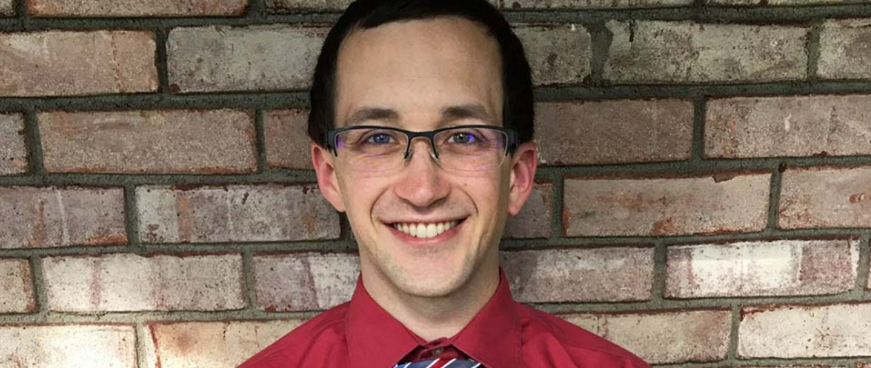 A young man wearing glasses and a red button down shirt smiles against a brick wall.