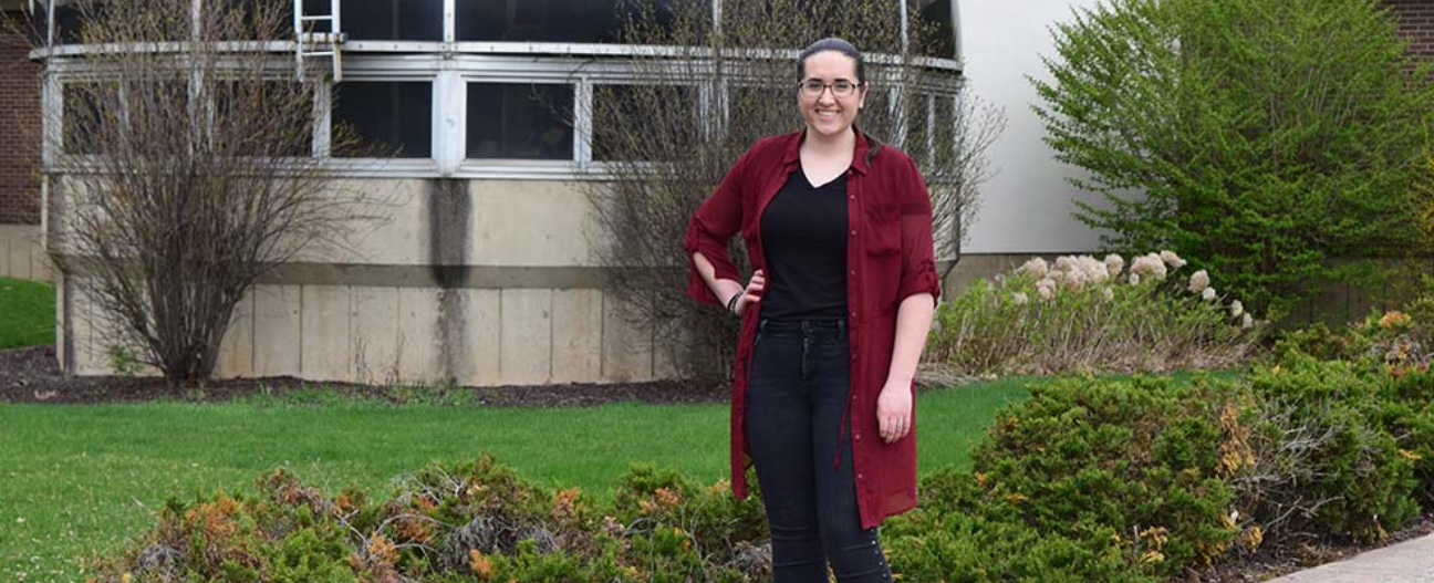 A woman wearing glasses, a black t-shirt, jeans and a red duster poses with her hand on her hip in front of a greenhouse on the LCCC campus.