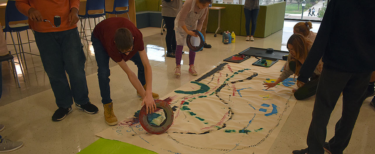 Students use materials to paint on a large piece of paper on a classroom floor.
