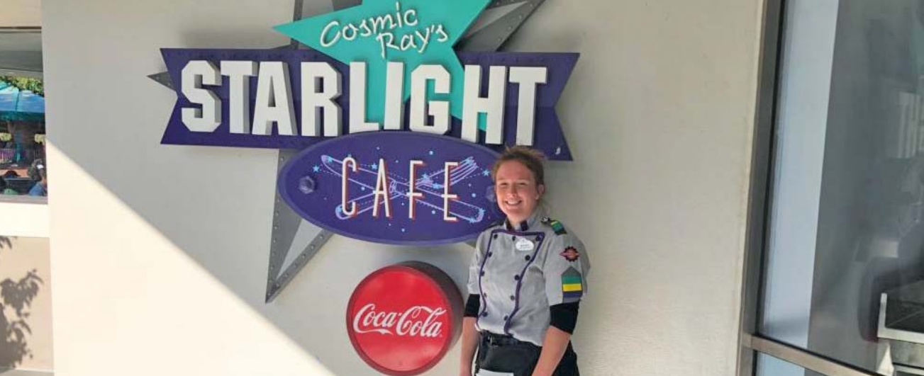 A woman in a work uniform poses in front of a sign that reads "Cosmic Ray's Starlight Cafe"