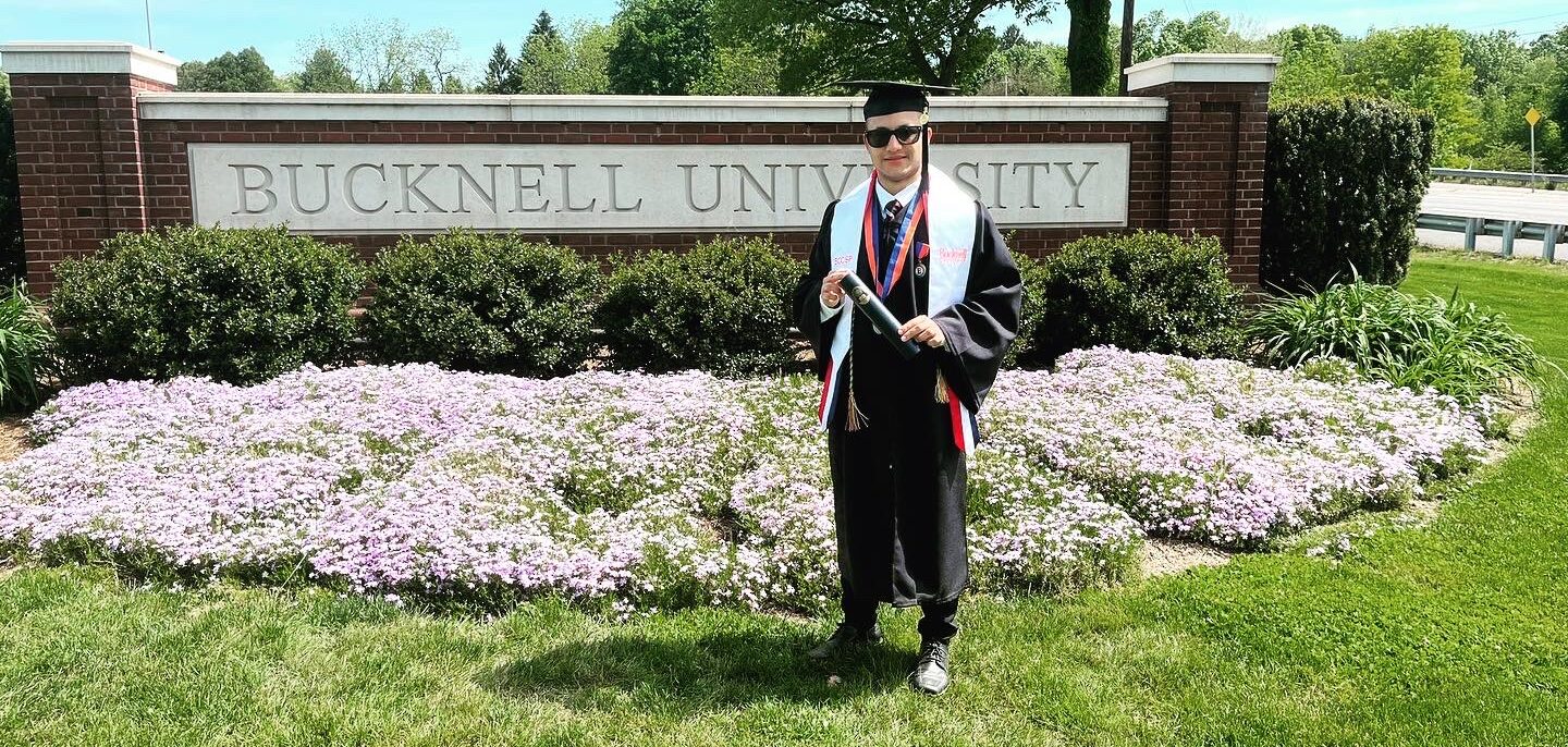 A graduate holding a diploma poses on a sunny day in front of the Bucknell University main campus sign.