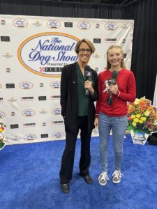 Two people with microphones stand in front of a wall with the National Dog Show logo on it.