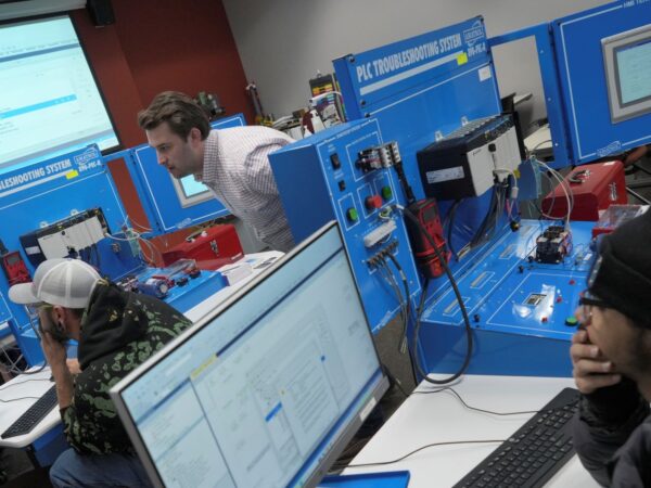 Two students sit at computer desks facing monitors displaying software interfaces in a technical classroom or lab setting. In the foreground, a student wearing a black beanie and a winter coat looks at a desktop screen. In the background, another student in a baseball cap works at a computer while an instructor in a patterned shirt leans over to inspect a large blue "PLC Troubleshooting System" training station with wired components and control panels. A projection screen displaying computer code or software is visible in the upper-left background.