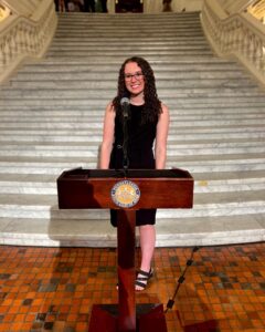 A young woman with dark brown curly hair and glasses stands at a wooden podium in front of a large staircase.