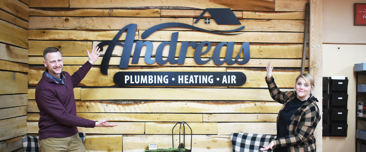 A man and woman stand on either side of a wall that features a logo that reads "Andreas Plumbing Heating Air"
