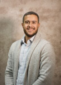 A man wearing a checkered shirt and cardigan poses in front of a photography backdrop.