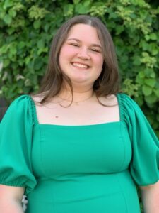 A woman with long brown hair and wearing a green dress, smiles in front of greenery.