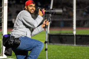 A man in a gray long sleeve shirt, jeans and red hat kneels to take a photo on a sports field.