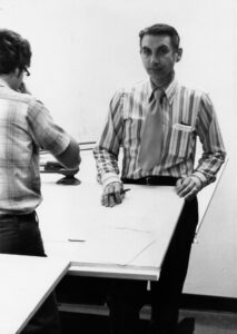 An old black and white photo shows a young Clifford Miller at a drafting table with another person whose back is toward the camera.