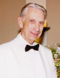An older man in a white tuxedo jacket smiles at the camera.