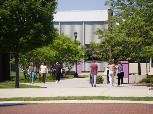 A sunny, wide-angle shot of a brick and concrete walkway on a college campus, lined with green trees and lampposts. In the background, a few small groups of students are seen walking toward and away from a large, modern academic building with light-colored siding.