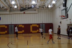 Six men's basketball players on a basketball court as one player shoots from the 3 point line. The compressed bleachers spell CCC in gold lettering.