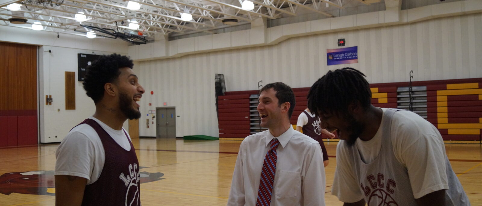 A man in a dress shirt and tie stands laughing with two two basketball players on a basketball court.