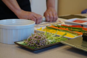 Shallow dishes filled with ingredients such as peppers, scallions, sprouts and fish are on a table while two hands roll a sushi mat. 