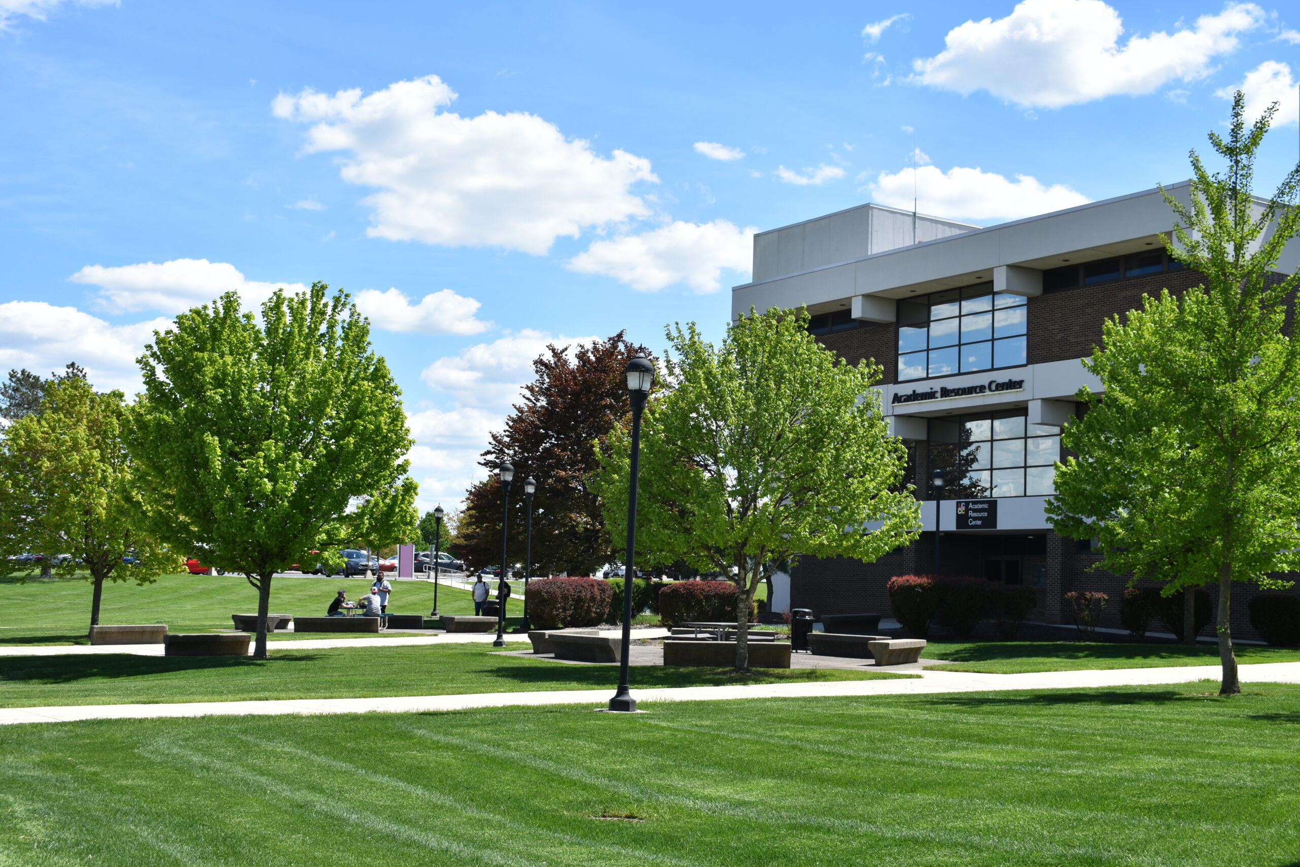 A sunlit outdoor view of the Lehigh Carbon Community College (LCCC) campus featuring green lawn areas, manicured trees, walkways, and a modern multi-story college building under a blue sky with scattered clouds.