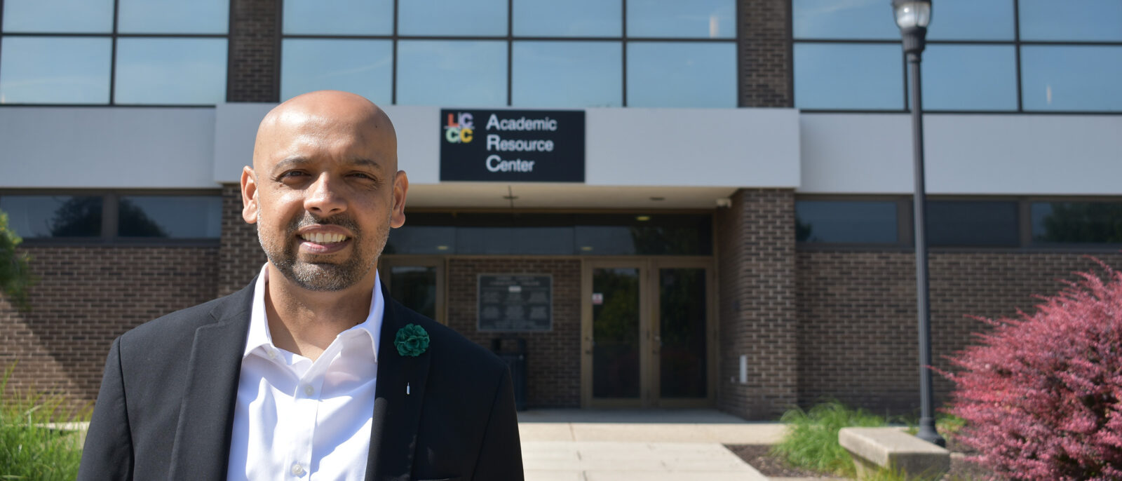 A man in a white dress shirt and black suit jacket stands outside the Academic Resource Center on the LCCC main campus.