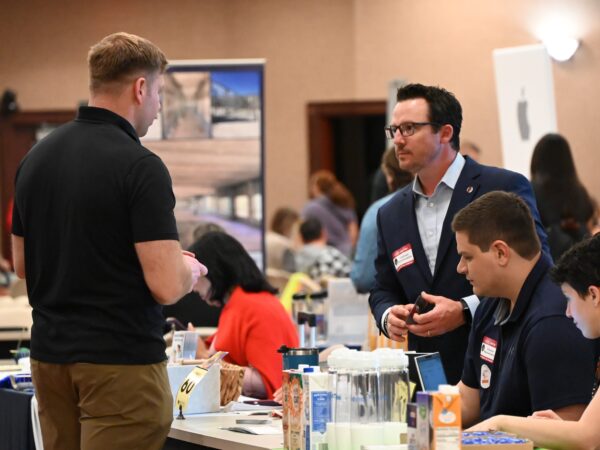 Professional networking event in a large hall where a representative in a navy blazer converses with attendees standing and seated around information tables.