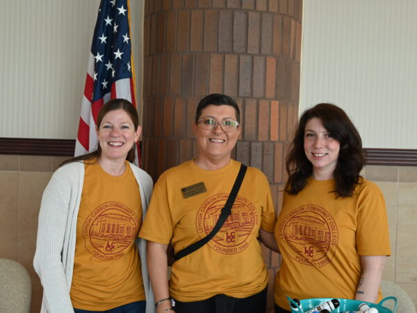 Three people standing in a row indoors, wearing matching orange t-shirts. The person on the left also wears a white cardigan. They are posing in front of a brick pillar and a wall with an American flag visible in the background.