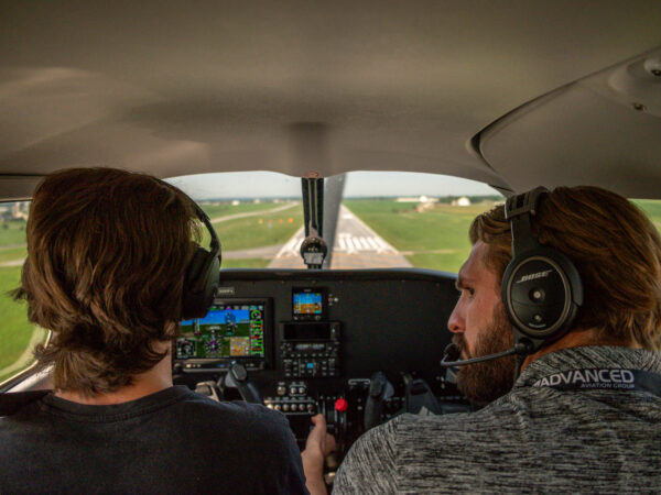 A view from behind the seats inside the cockpit of a small aircraft. Two people wearing aviation headsets sit in the pilot and co-pilot seats, facing forward toward the windshield, which shows an approach to a paved landing runway surrounded by green fields.