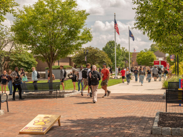 Students walking outdoors on a brick pathway on the Lehigh Carbon Community College (LCCC) campus, with trees, benches, flagpoles, and a cornhole game board visible in the courtyard.