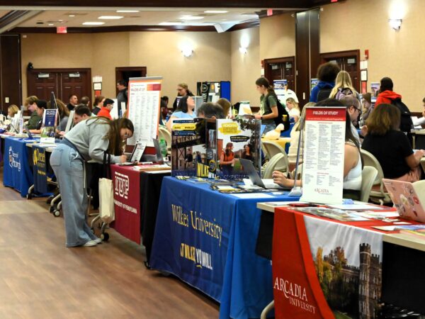 A college transfer fair taking place in a large indoor event hall, with tables set up by various universities including Arcadia University, Wilkes University, Drexel University, and IUP (Indiana University of Pennsylvania). Representatives and students interact across the tables, which display informational banners, brochures, and laptops.