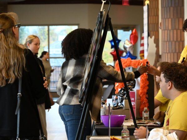 Students and visitors gathered around informational tables inside a school building during a event. On the right, representatives in yellow shirts sit behind tables displayed with flyers, banners, and decorations, while visitors on the left browse posters on an easel and interact with the hosts.