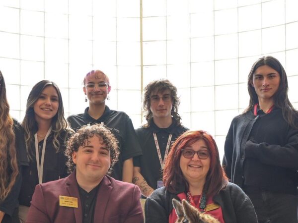 A group portrait of nine people and a service dog in front of a large grid window with soft, natural lighting. Two people sit on a patterned armchair in the foreground: a person in a maroon blazer on the left and a person with red hair and glasses on the right, petting a German Shepherd wearing a working harness. Seven people stand behind them wearing matching black polo shirts with lanyards around their necks.