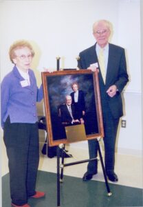 A man and woman stand on either side of a framed portrait of themselves.