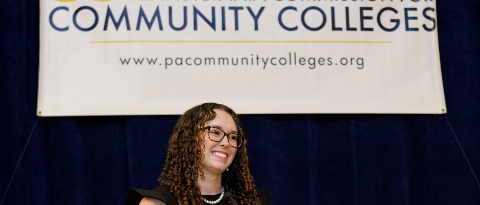 A young woman with dark brown curly hair and glasses stands smiling in front of a large banner that reads "Pennsylvania Commission for Community Colleges"