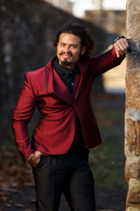 Alani Jimenez leans one arm against a tree while looking at the camera. He is dressed in a red suit jacket. 