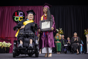 A faculty member in a motorized chair poses next to a student in commencement regalia holding a framed award on a stage with the LCCC logo on a curtain in the background.