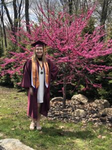 A graduate wearing commencement regalia stands in front of a red bud tree.