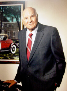 Man in a suit stands next to a framed photo of a vehicle that is partially in the photo.